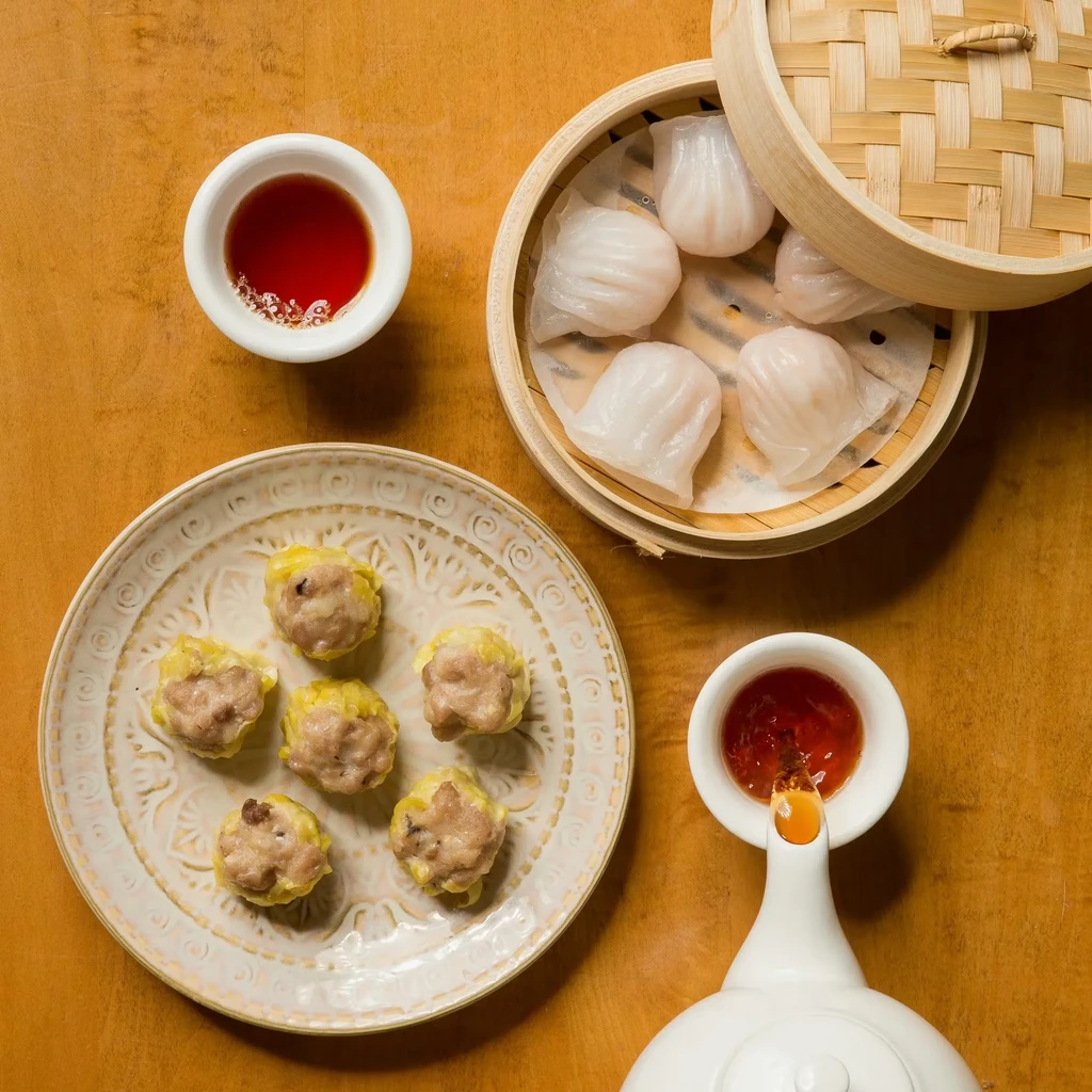 Dim sum dishes: Siu Mai, Har Gow, and sauces on a wooden surface.