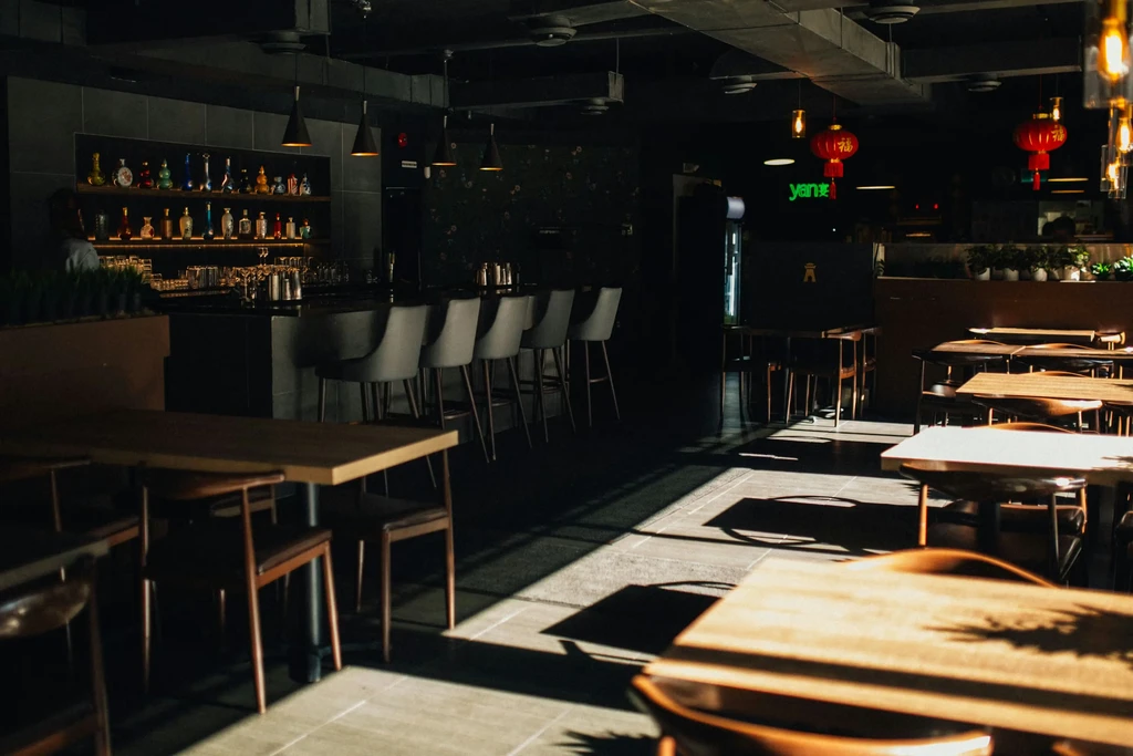 Dark restaurant interior with a bar, sunlit tables, and Chinese lanterns.