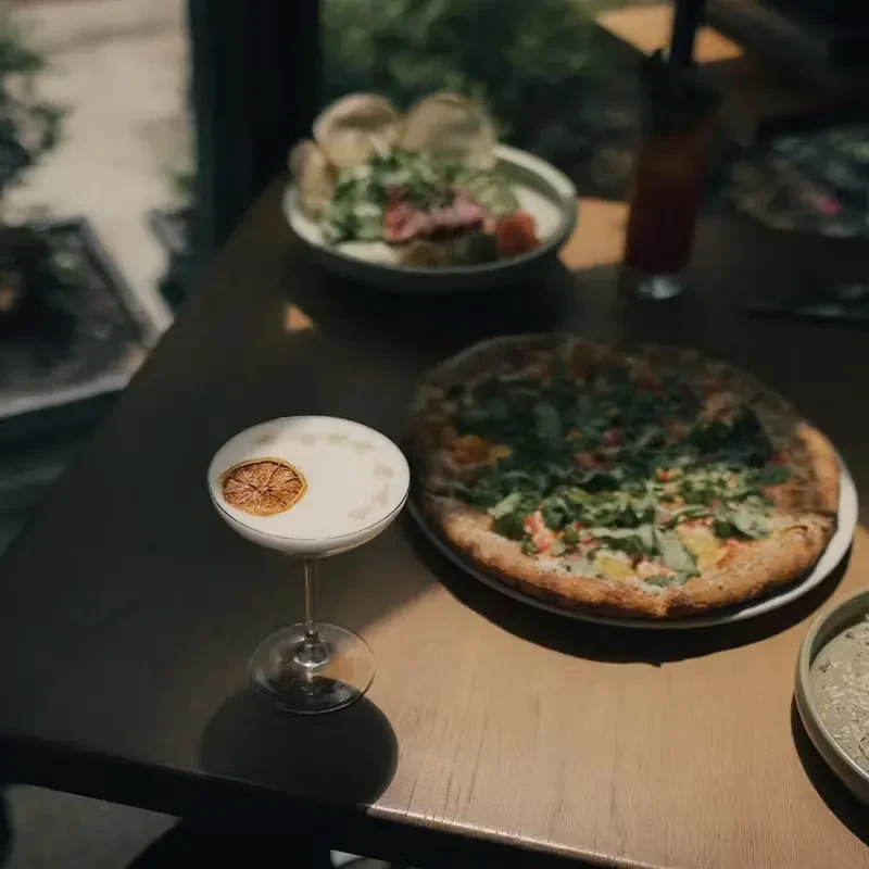Frothy cocktail, arugula pizza, and salad on a sunlit dining table.