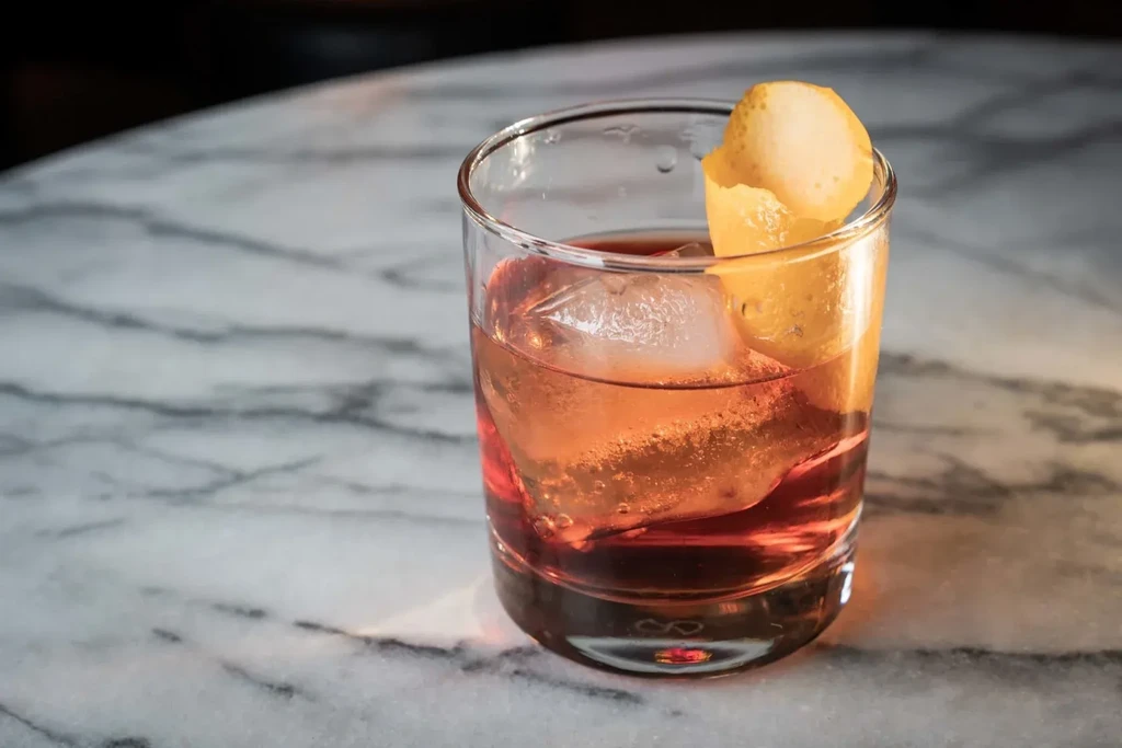 Reddish-orange cocktail in rocks glass with large ice and citrus peel.
