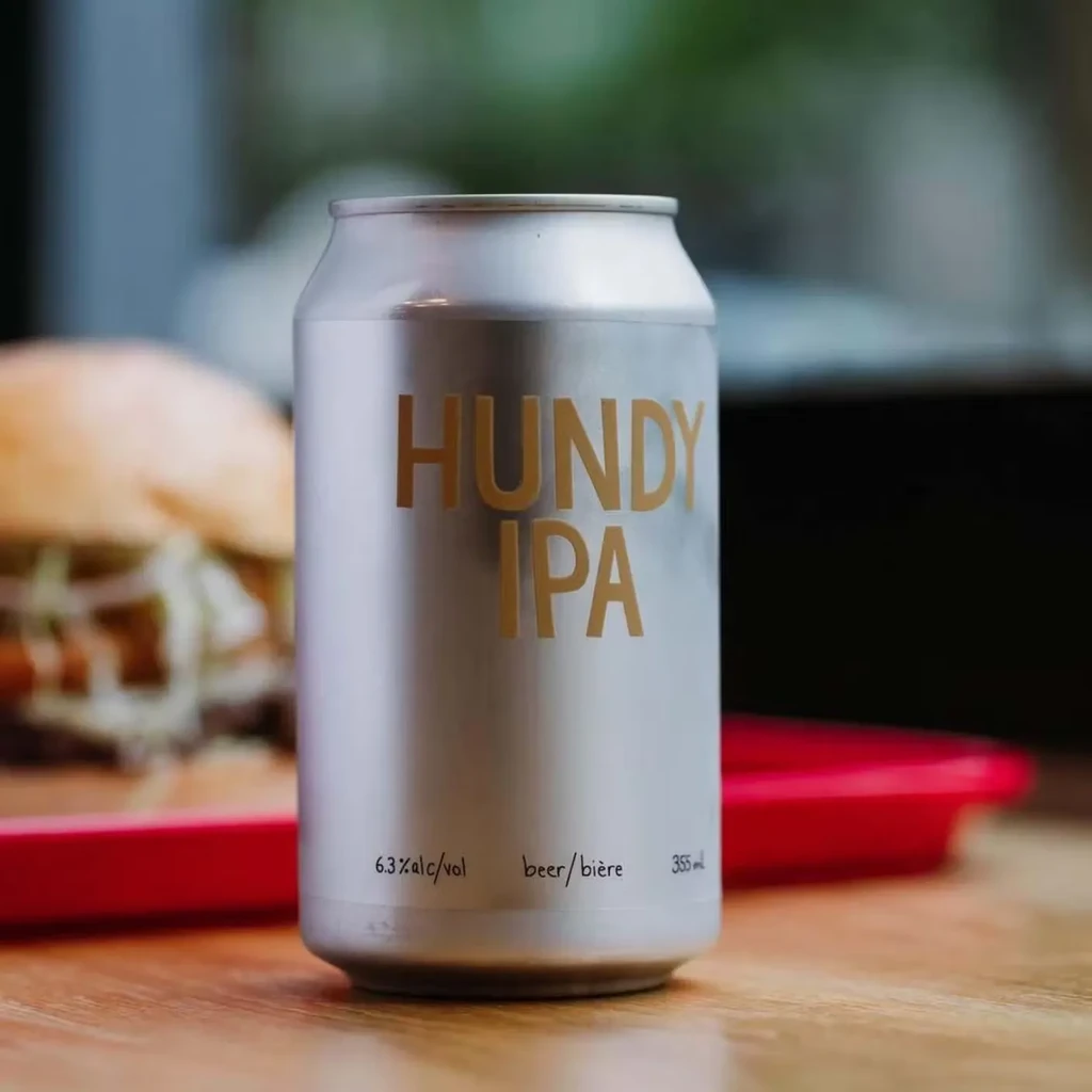 Silver HUNDY IPA beer can with a blurred hamburger in the background.