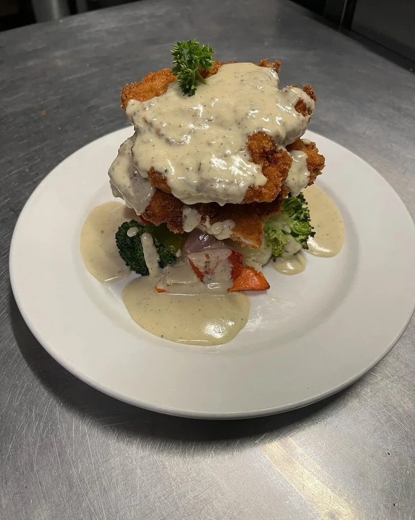 Stacked fried protein with creamy sauce, colorful vegetables, and parsley.
