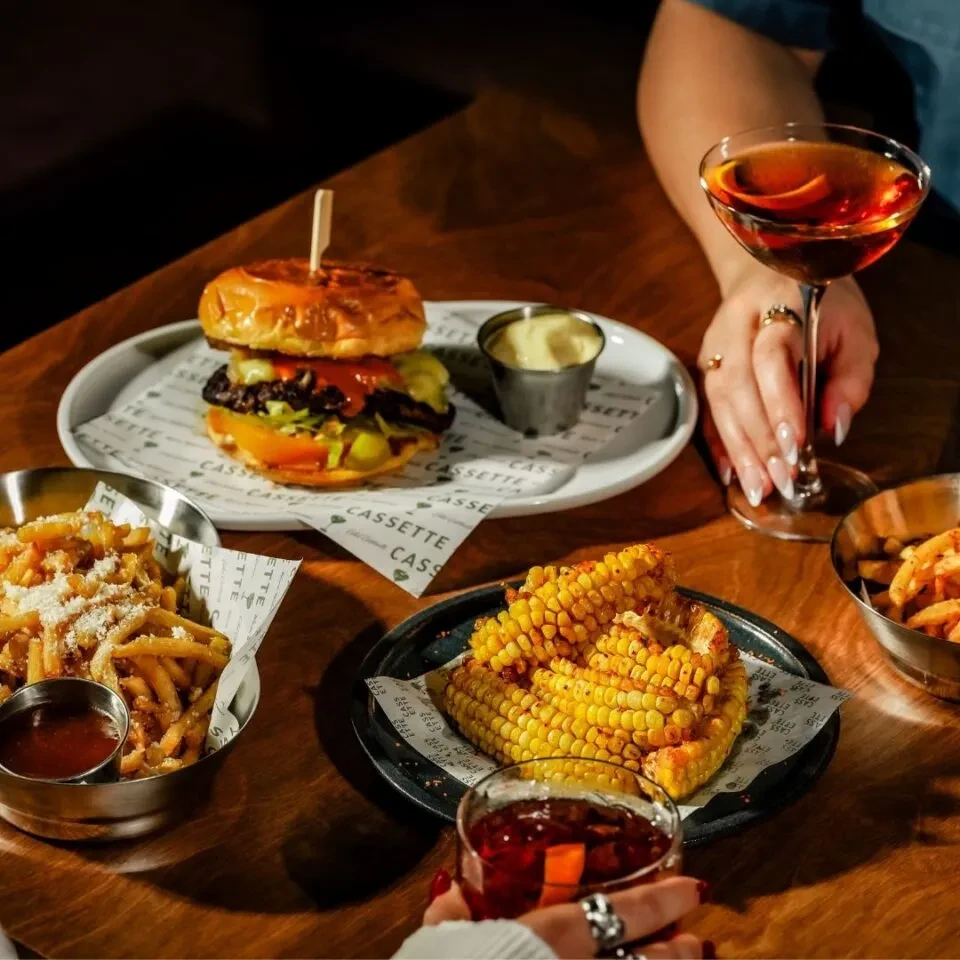 Gourmet burger, cheesy fries, grilled corn, and two cocktails on a table.