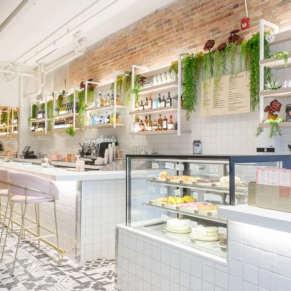 Bright modern cafe: white counter, pink stools, pastry case, plants.
