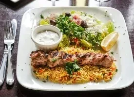 Souvlaki skewer, seasoned rice, salad, tzatziki, and lemon on a plate.