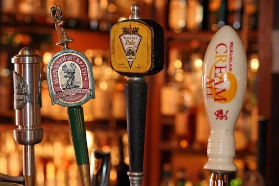 Four branded beer tap handles lined up in a well-stocked bar.