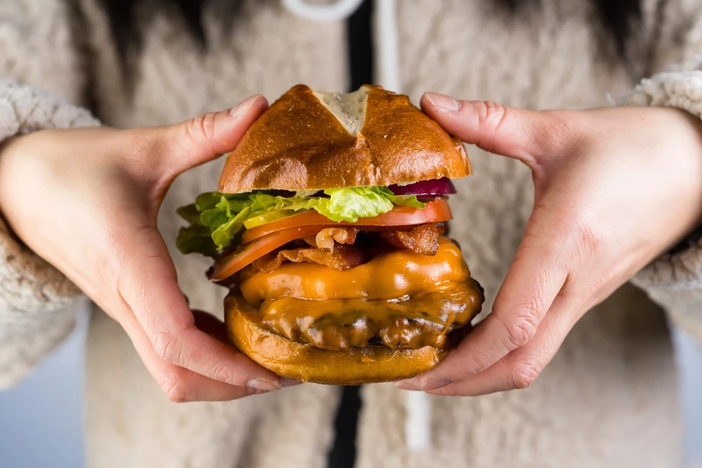 Burger with pretzel bun, melted cheese, bacon, and vegetables, held by hands.