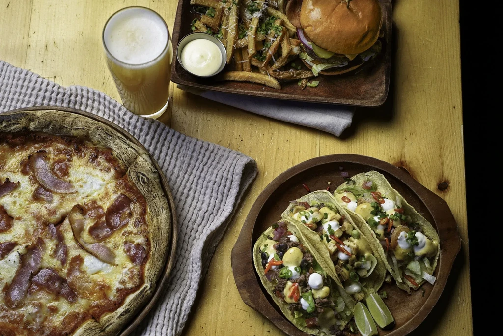 Overhead shot of pizza, beer, burger, fries, and tacos on a rustic table.