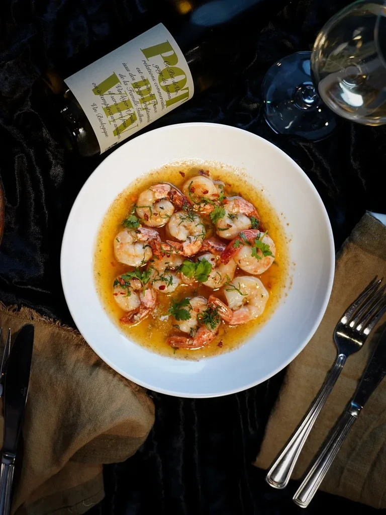 Overhead shot: bowl of shrimp with herbs, wine bottle, glass, and cutlery.