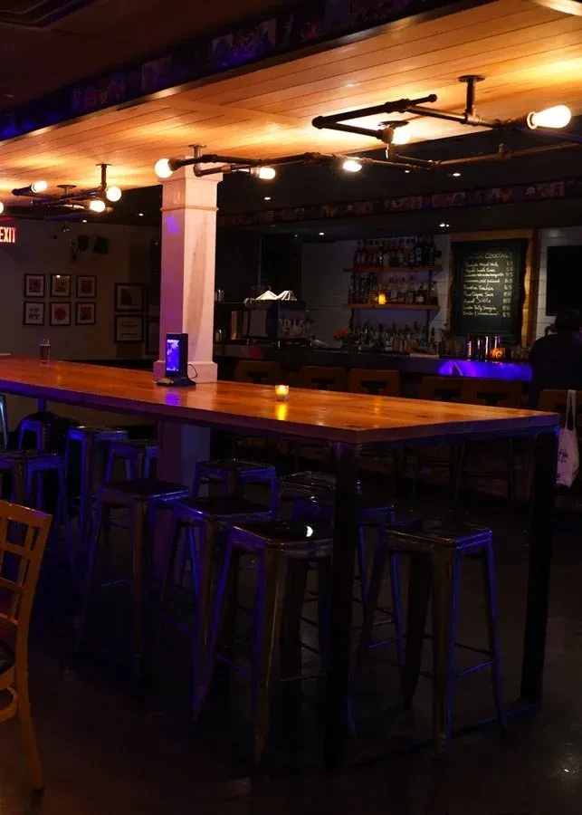 Dimly lit industrial bar with long communal table and stocked bar.