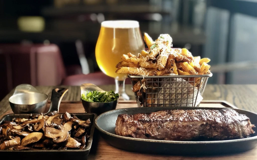 Steak, sautéed mushrooms, cheesy fries, and beer on a wooden table.