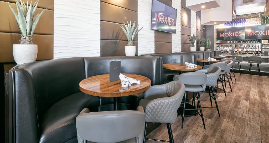 Modern upscale restaurant interior with booth seating, bar, and MOXIES TVs.