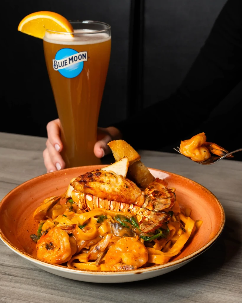 Hand forks shrimp from pasta with lobster, beside Blue Moon beer.
