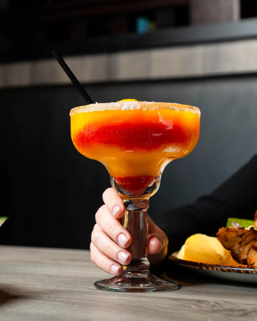Hand holds vibrant layered frozen margarita with frosted rim.
