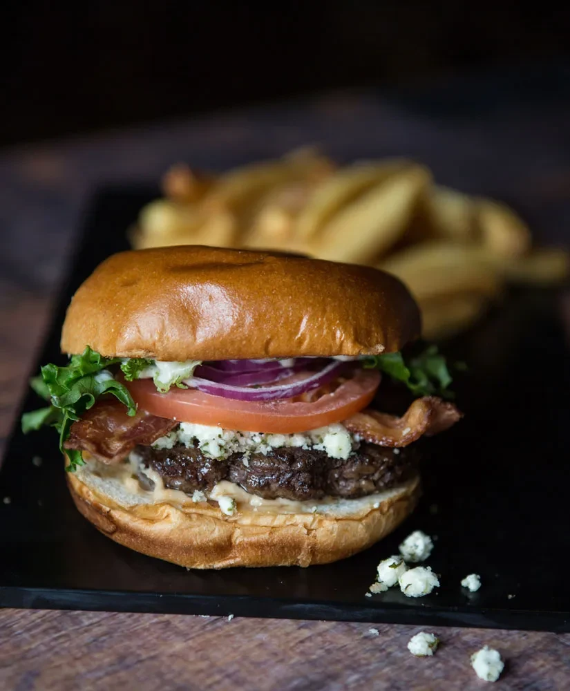 Close-up of a gourmet burger with blue cheese, bacon, and fries.