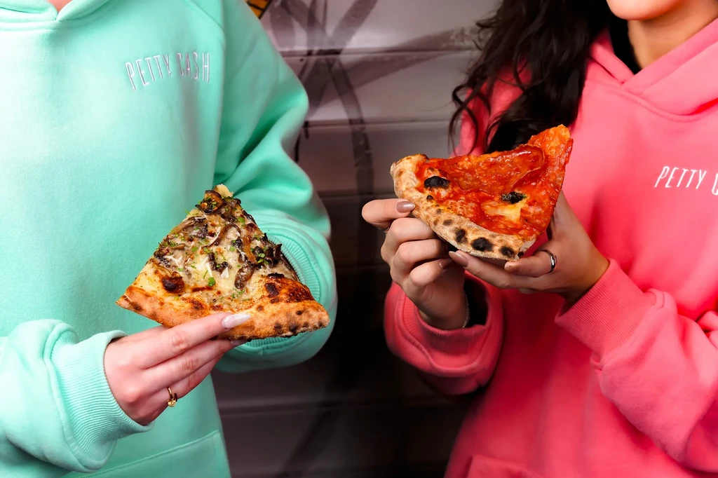 Two people in "PETTY CASH" hoodies holding mushroom and pepperoni pizza slices.