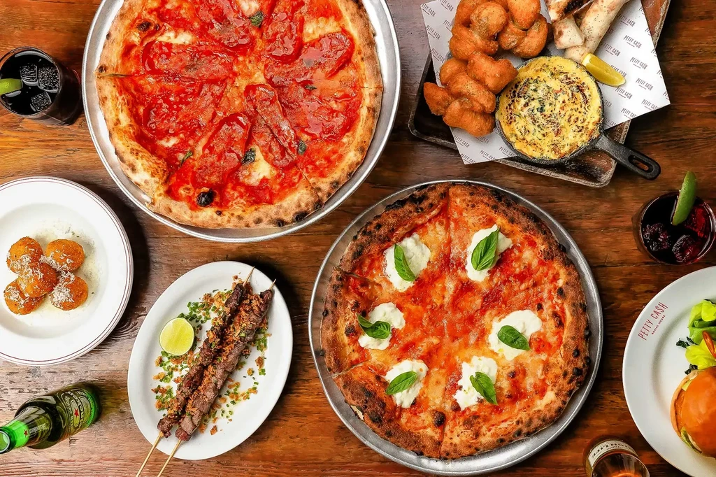 Overhead shot of a diverse food spread with pizzas, skewers, and drinks.