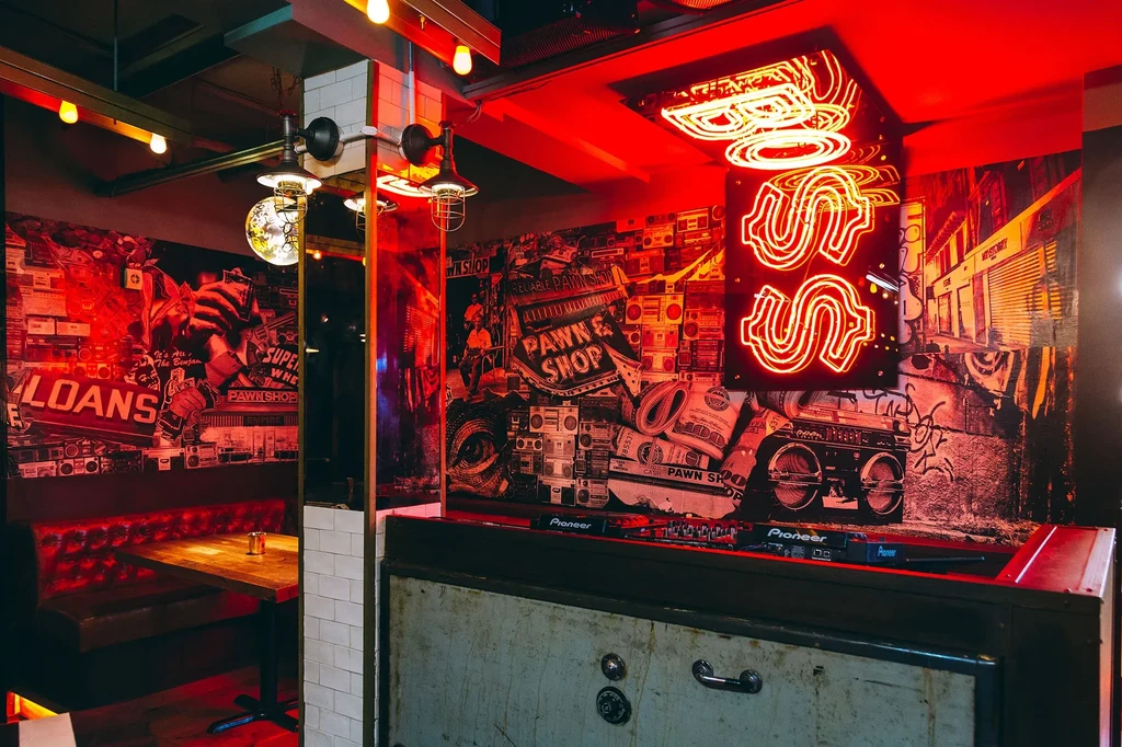 Red-lit urban bar with collage murals, neon dollar sign, and DJ equipment.