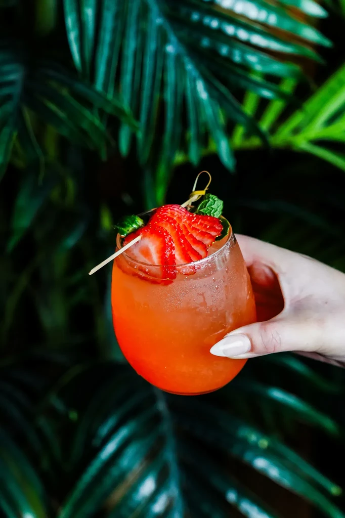 Orange-red cocktail with strawberry and mint, held against tropical foliage.