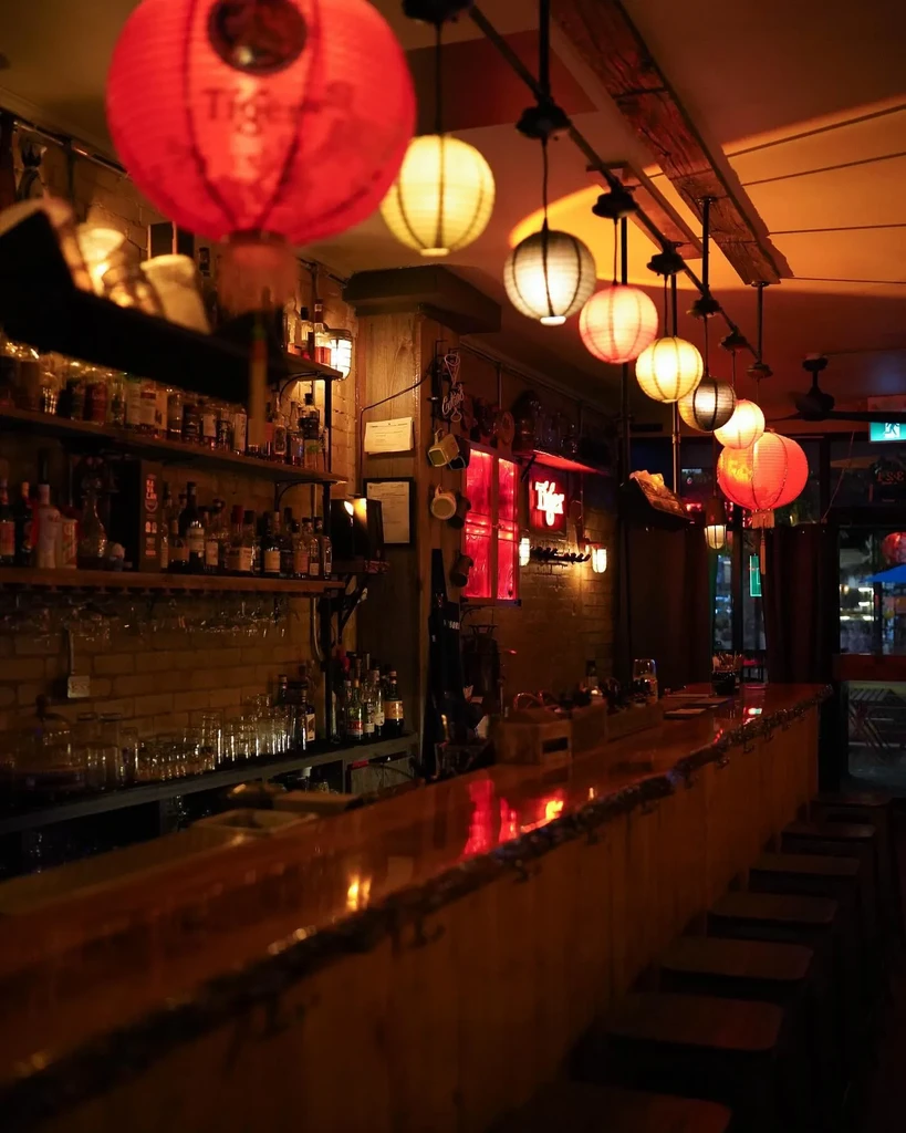 Dimly lit bar with wooden counter, shelves of bottles, and Tiger lanterns.