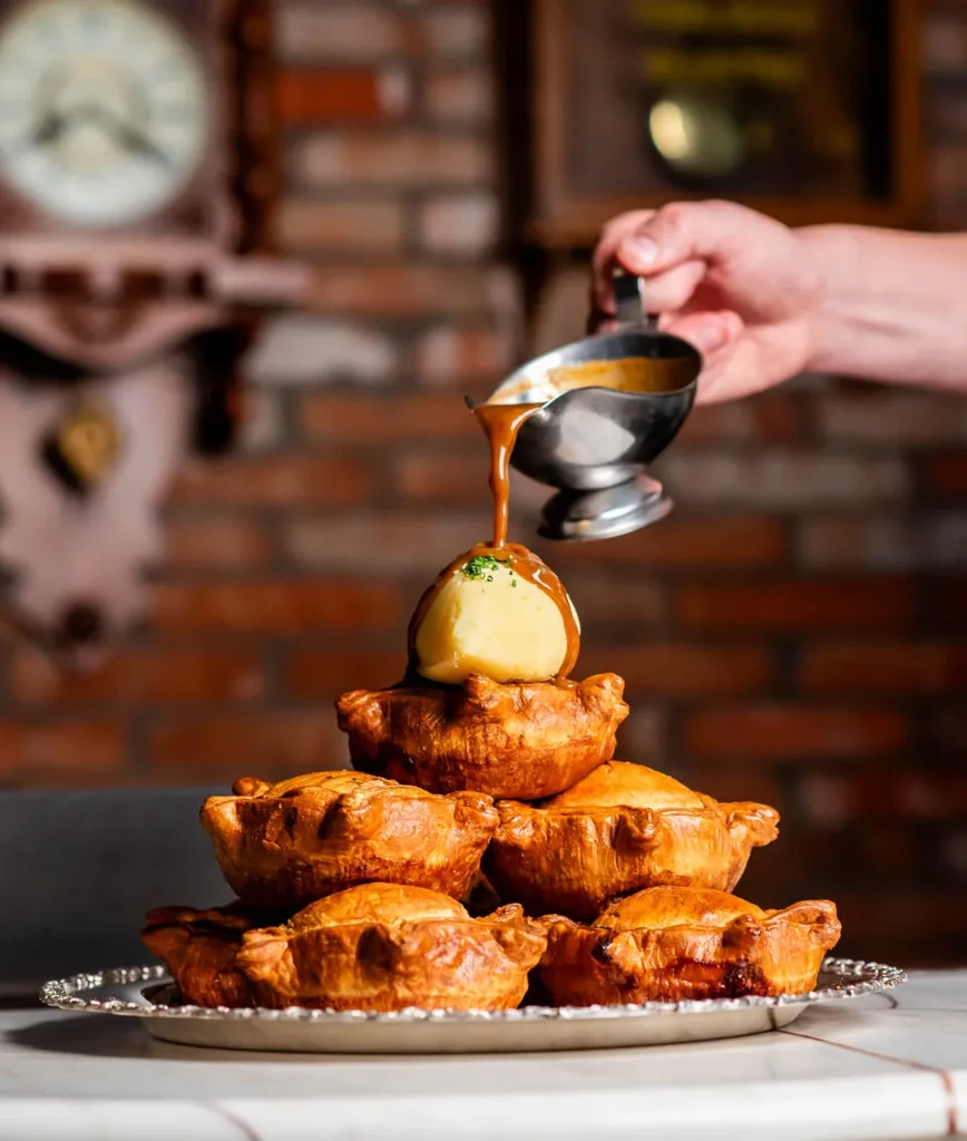 Hand pours gravy onto chive mashed potatoes atop stacked meat pies.