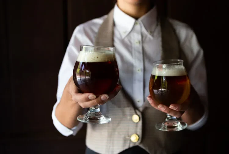 Person in vest holding two stemmed glasses of dark, foamy beer.
