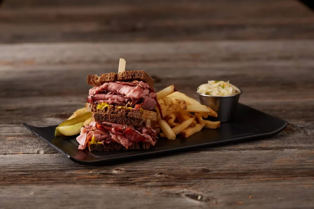 Stacked deli sandwich, fries, coleslaw, and pickle on black plate.