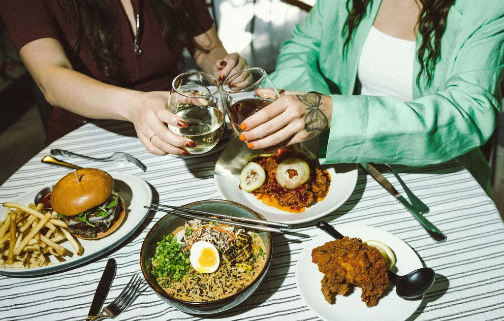 Overhead table view: burger, ramen, fried chicken, hands toasting glasses.