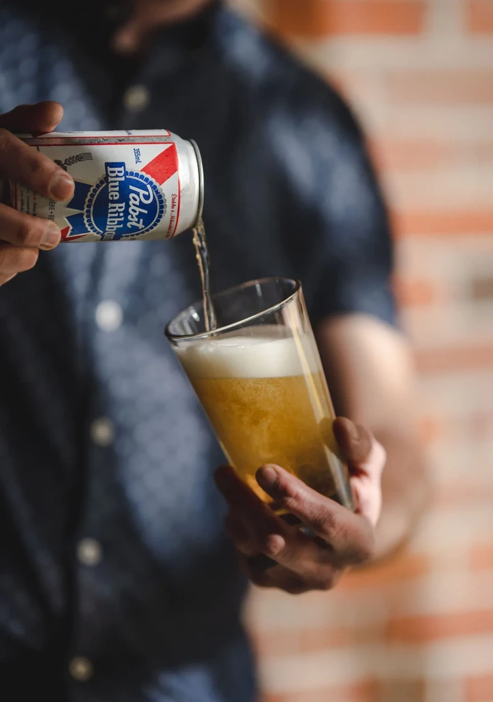 Close-up of hands pouring Pabst Blue Ribbon beer into a glass.