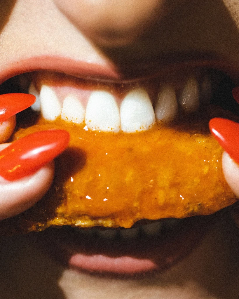 Mouth with red lipstick biting into an orange sauced chicken wing.