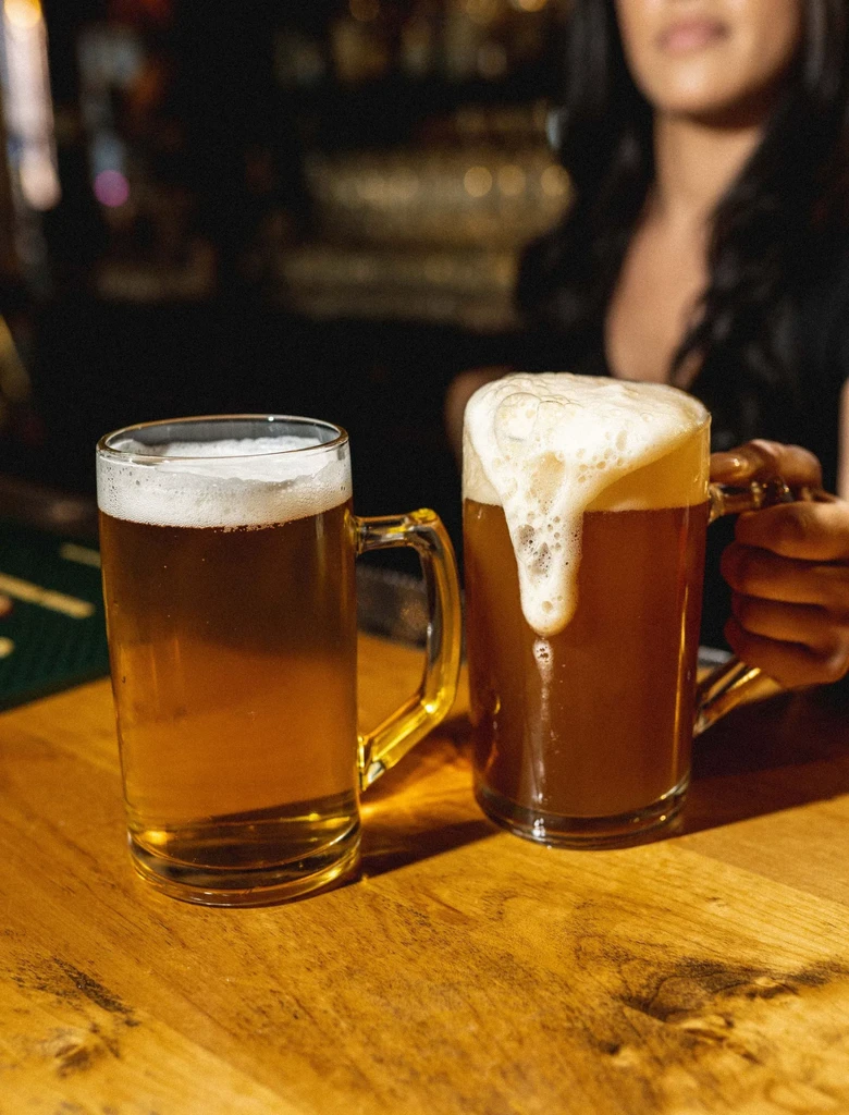Two amber beers, one overflowing, on a dim wooden bar.