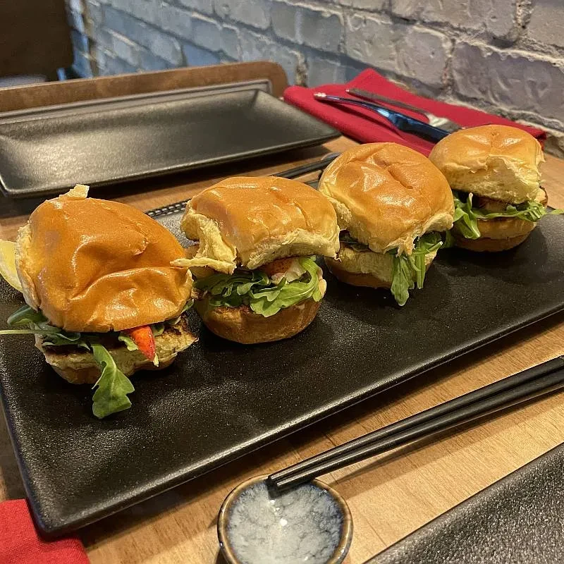 Four sliders with lettuce and reddish filling on a black plate.
