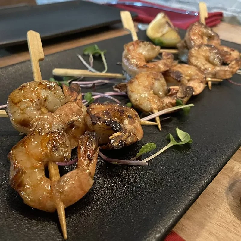 Two grilled shrimp skewers with microgreens and lime wedge on a black slate.