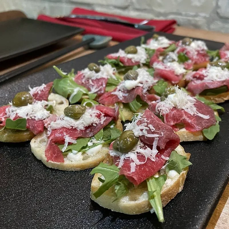 Crostini appetizers with meat, arugula, parmesan, and capers on a dark platter.