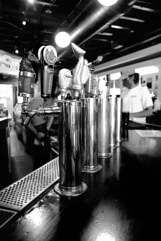 Black and white photo of six shiny beer taps on a bar counter.