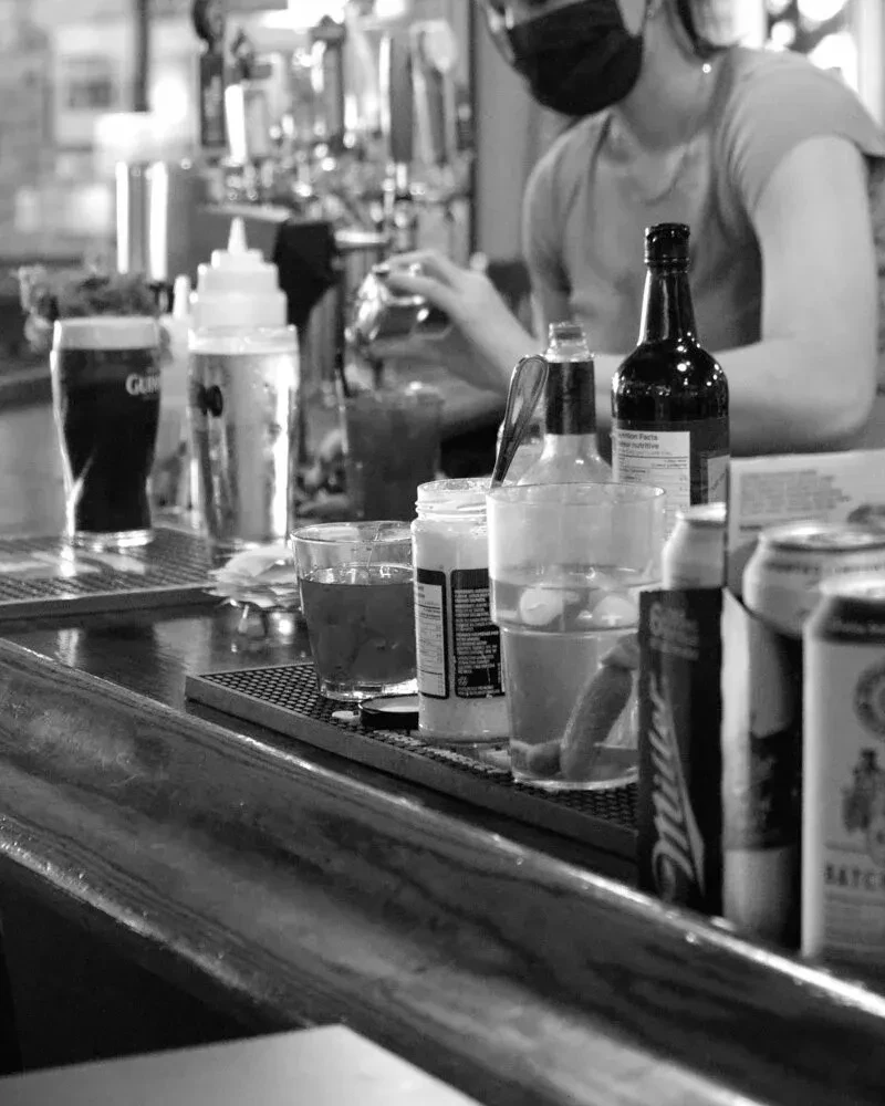 Masked bartender preparing drinks behind a busy bar counter. Black and white.