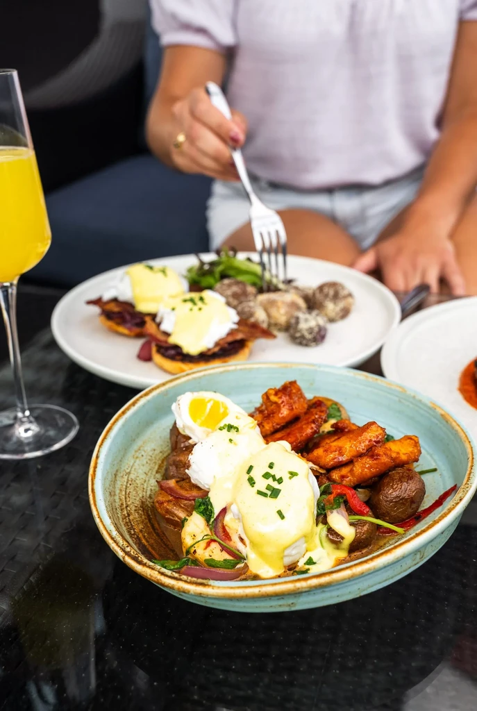 Bowl of poached eggs, potatoes, and Eggs Benedict with a drink.