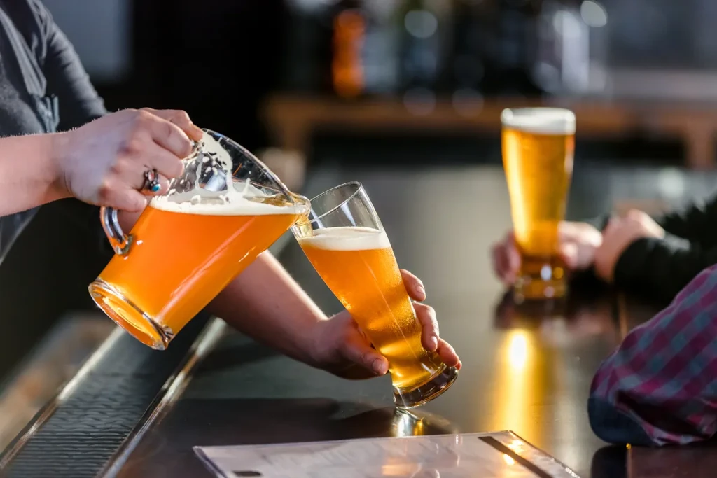 Hands pouring frothy golden beer from a pitcher into a tall glass.