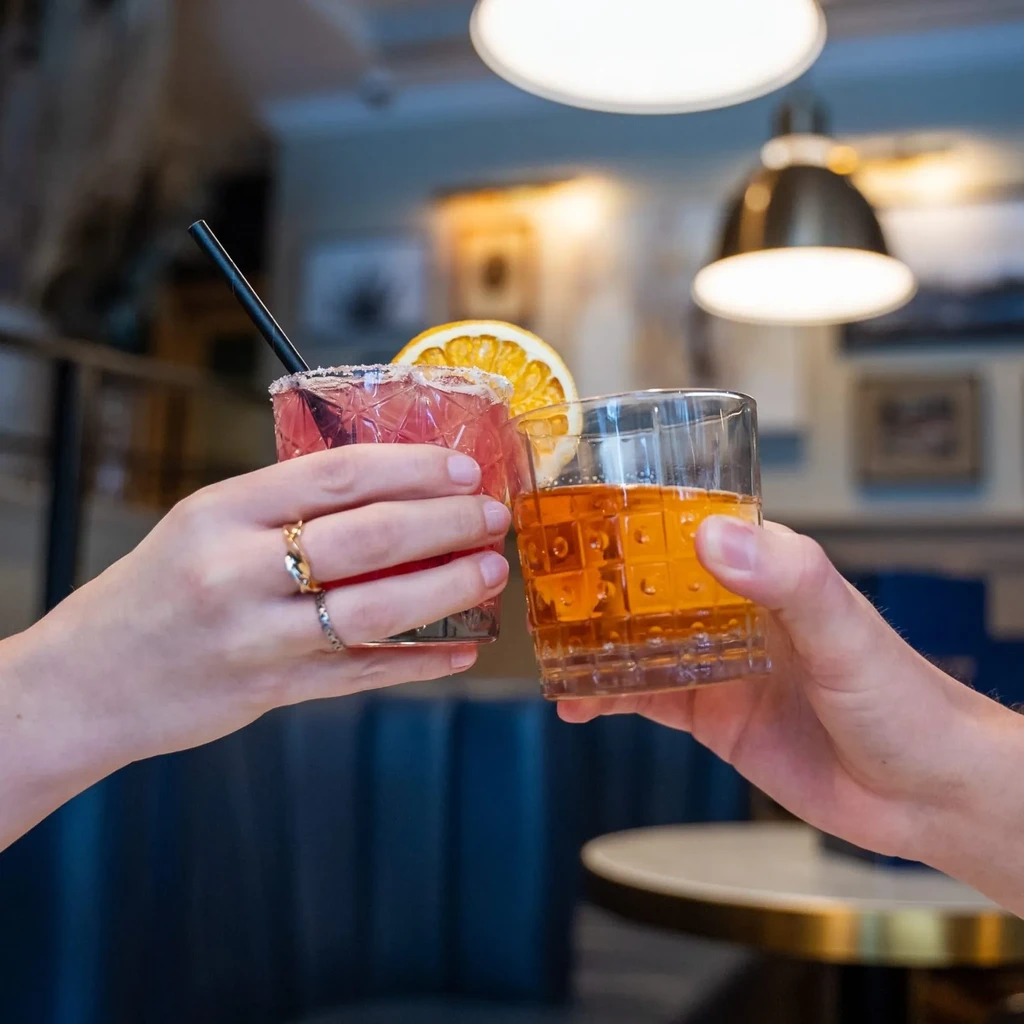 Close-up of two hands toasting with pink and amber cocktails.