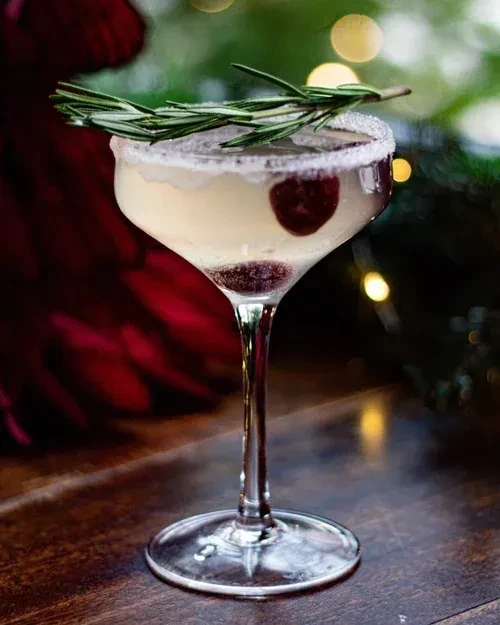 Cocktail in coupe glass, light liquid, sugar rim, red berries, rosemary.