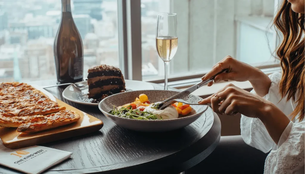 Person cutting salad, cake, and flatbread at a city view restaurant.