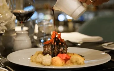 Hand pouring sauce over a plated meat and vegetable dish.