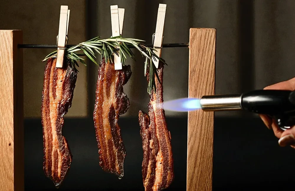 Three crispy bacon strips with rosemary, suspended and torched.
