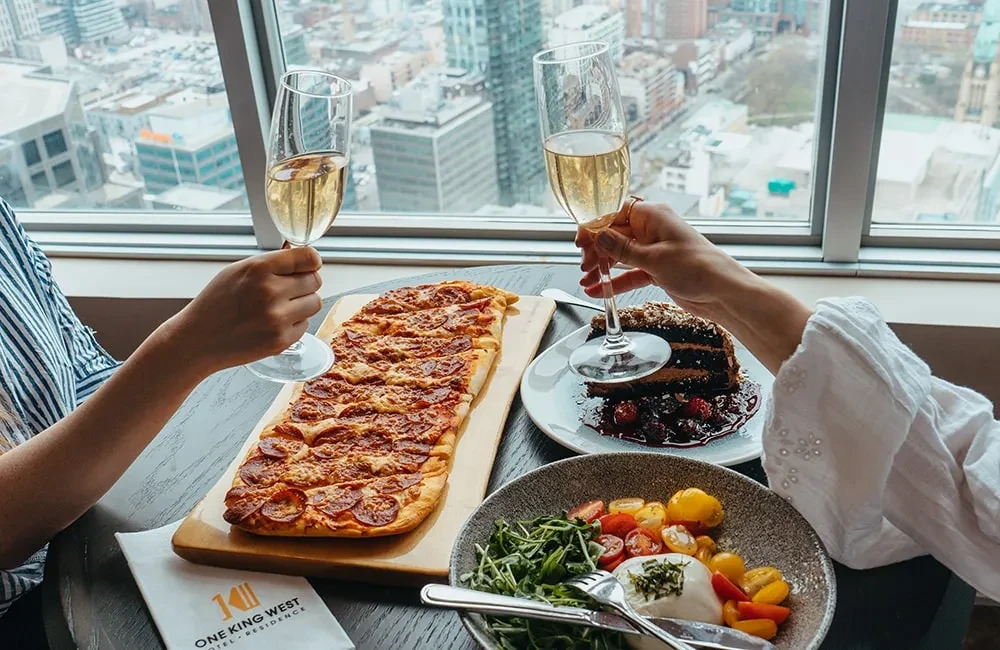 Hands toast champagne over pizza, cake, salad, and city view.