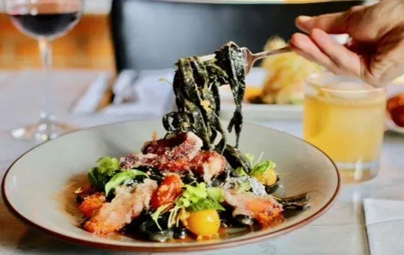 Close-up: hand forks dark pasta with seafood, tomatoes, cheese.
