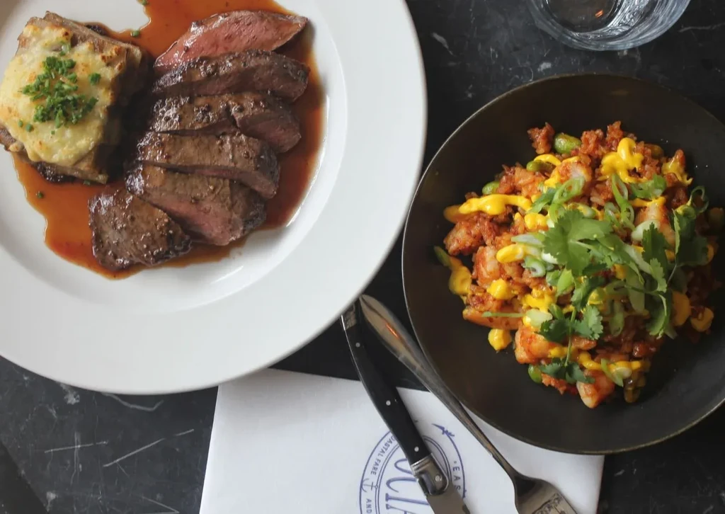 Sliced meat with potato gratin and shrimp rice bowl on dark table.
