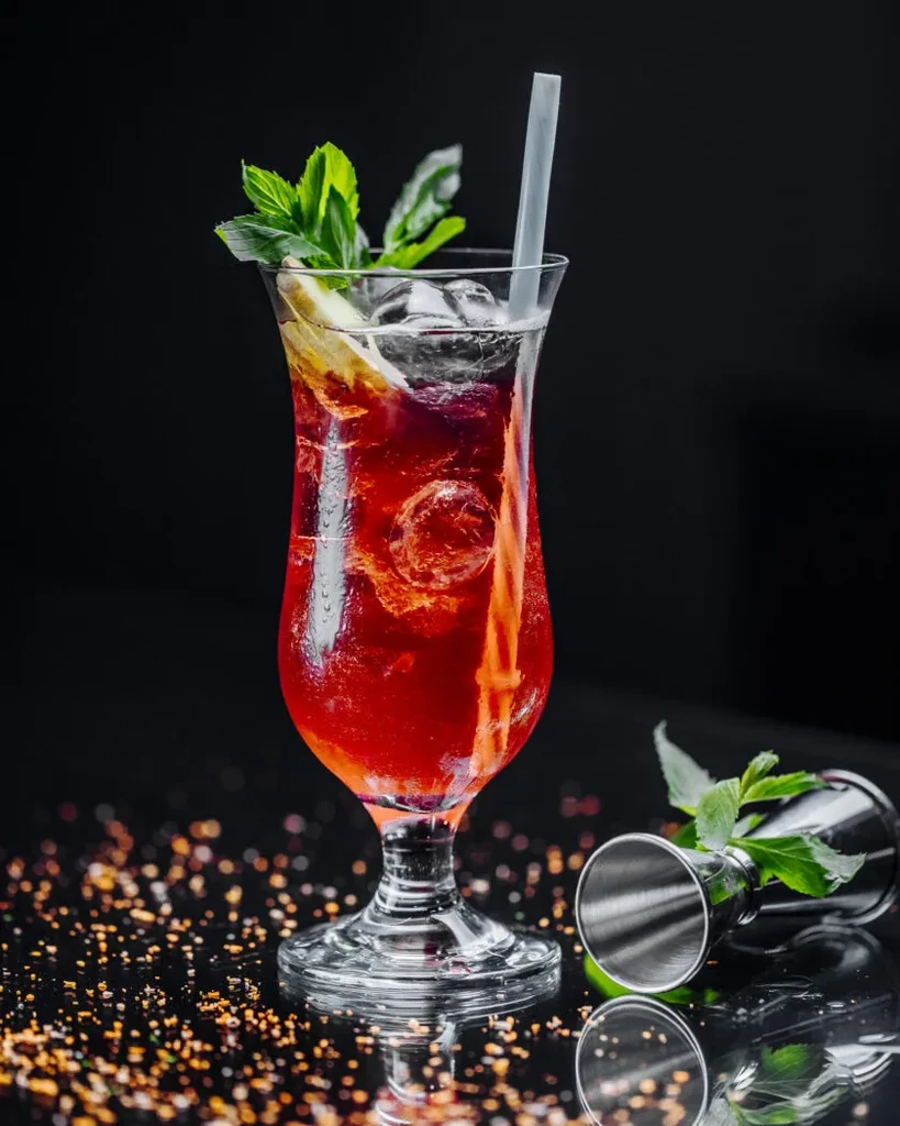 Red hurricane cocktail with mint, citrus, and ice on glittery surface.