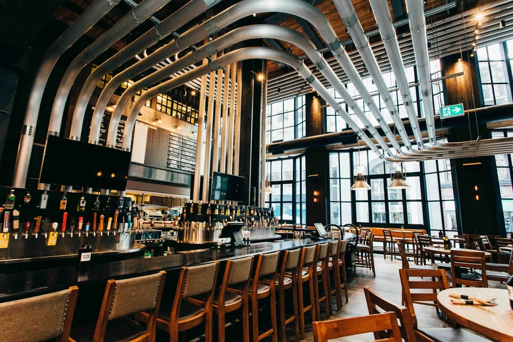 Industrial restaurant with curved pipes, long bar, and tall windows.