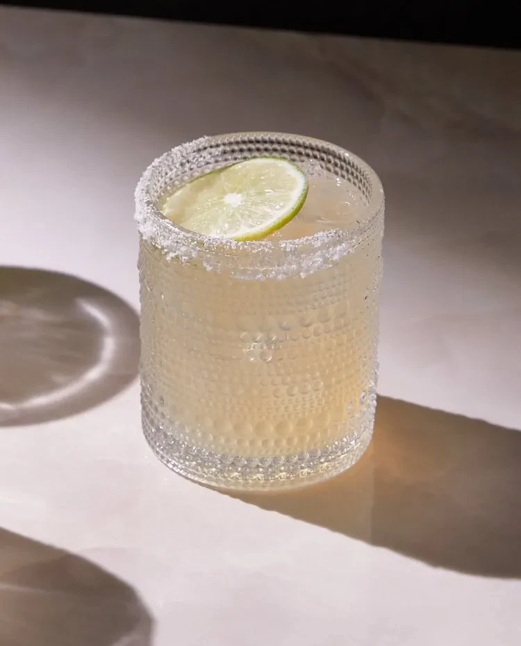 Pale yellow cocktail in a textured, salt-rimmed glass with lime.