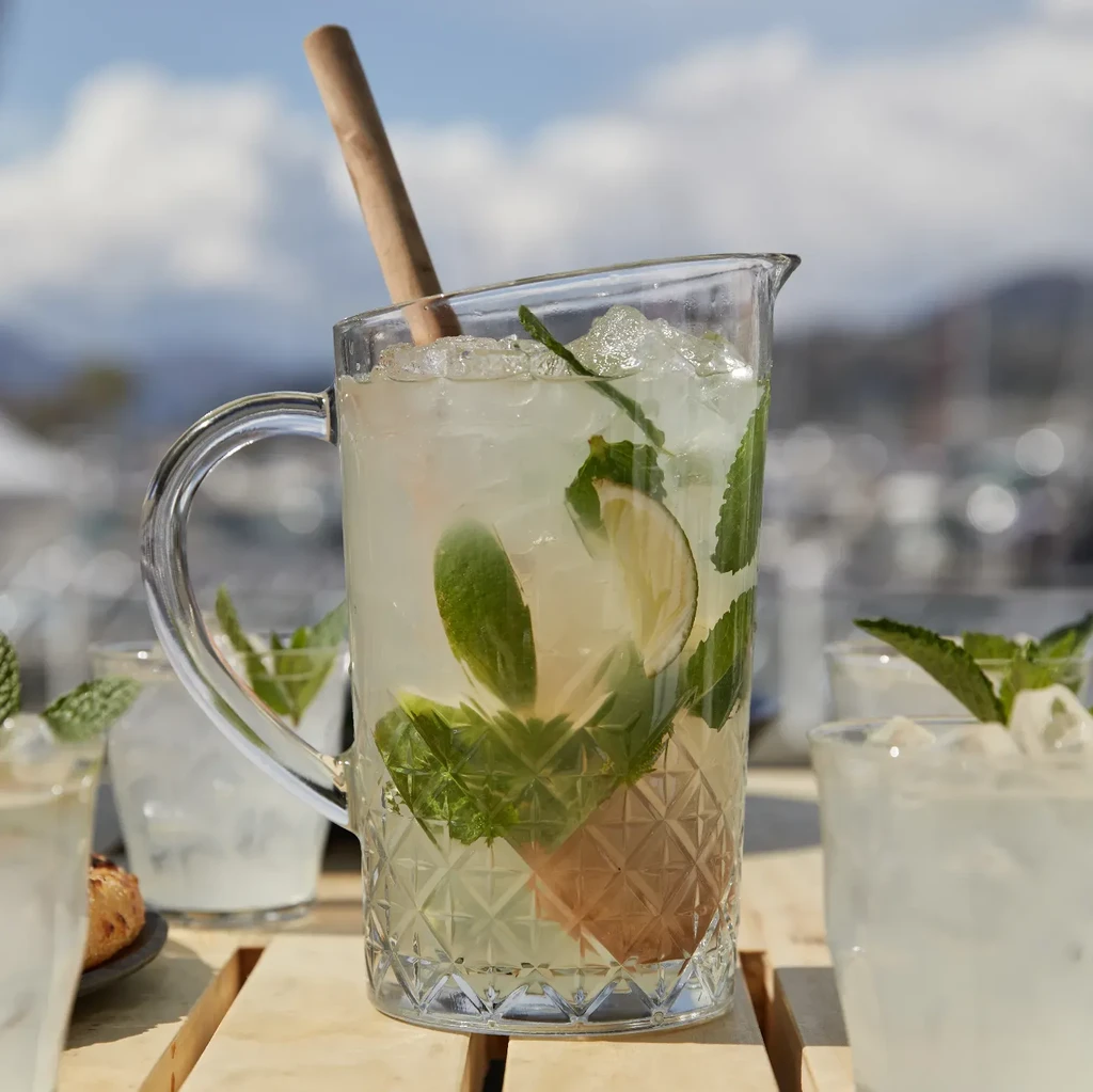 Large glass pitcher of mint lime drink outdoors by waterfront.